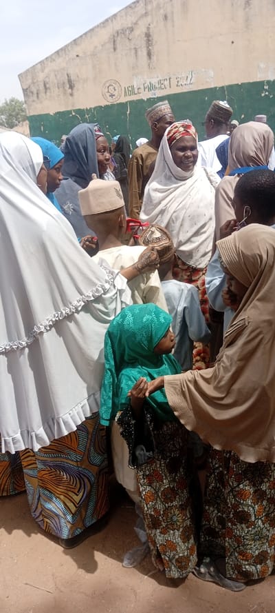 A crowded outdoor scene in Sani Mai Nagge, Nigeria, depicts approximately 10-15 individuals, predominantly women and children, gathered on dry, light brown earth under bright daylight. In the background, a weathered concrete wall, painted green on its lower section and light beige on its upper, displays the stenciled text "AGILE PROJECT 2022" alongside a circular logo featuring a stylized female figure.
In the mid-ground, a woman with prominent henna decoration on her left hand (partially obscured) is interacting with a young boy wearing a light beige cap. Close to this interaction, several red, spherical objects, resembling fruit, are visible in people's hands. To the right, another woman, wearing a beige head covering and patterned skirt, engages with a young girl in a teal head covering, their hands appearing to touch or exchange an item.
The individuals are dressed in various traditional attires, including multiple women wearing head coverings in white, blue, teal, and darker shades, as well as patterned skirts or wraps. At least one adult male is visible in the background, also wearing a patterned cap. The overall impression is a communal gathering or distribution event.