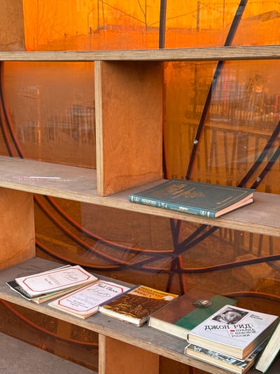 The image shows a bookshelf filled with books. The books are arranged in a disorganized manner on the shelves. The bookshelf is made of plywood and has an orange-colored back. Some of the books have Russian titles like "Джон Рид: Правда о Красной России" (John Reed: Truth About Red Russia). Another book is titled "Карл Маркс Ф.ЭНГЕЛЬС Манифест Коммунистической партии" (Karl Marx F. Engels Manifesto of the Communist Party). Another book is titled "Стихотворения" (Poems). There appears to be a reference to Karl Marx as well.
There is no way to determine the location as the image does not show anything to establish the location, even if the texts on the books suggest the location to be somewhere in Russia.