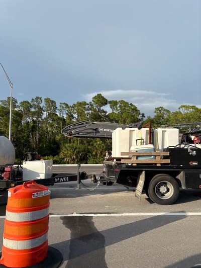 An outdoor, medium shot captures a flatbed truck with multiple water tanks and equipment parked on a road, likely for construction or maintenance work, under a cloudy sky. The truck has "W WIRTS" visible on a piece of equipment, and "WW01" on its side. In the foreground, an orange traffic barrel with reflective bands is positioned on the asphalt. The background features a dense line of tall, green trees. The scene is illuminated by natural light, casting shadows on the road.