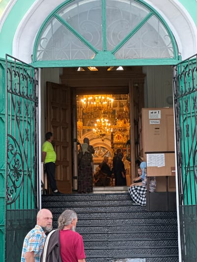 Captured from an oblique angle, the image shows a partial view into what appears to be the interior of a religious structure, possibly a church, in Diveyevo, Russia. The scene is framed by an open, decorative metal gate painted in a pale green hue, and a semi-circular window with ornate glasswork.
Inside, a large chandelier dominates the interior, reflecting light throughout the space. Several individuals are visible, including people in a line and one person lying down in the doorway. There are also objects such as boxes and containers within the room, adjacent to the line.
Outside, two men stand near the gate, partially obscuring the view. They are dressed in casual clothing and appear to be looking towards the interior of the building.