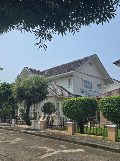 This image depicts a residential street scene in San Phra Net, Thailand, under a clear blue sky. The foreground features an asphalt road surface exhibiting discoloration and wear, bordered by a raised concrete curb. Overhanging dark green foliage from a tree canopy partially obscures the sky in the upper left quadrant.
In the midground, a two-story residential building with light-colored (cream/off-white) walls and a dark gray tiled roof dominates the view. The house features multiple gables, white trim, and white-framed windows, including a second-story balcony with white railings. A black metal picket fence with light-colored concrete pillars and beige bases delineates the property line from the street. Behind this fence, the meticulously maintained landscaping includes several round-pruned green bushes positioned between fence posts, as well as various trees and green grass within the yard.
Portions of other similarly styled houses or extensions are visible to the right of the primary dwelling. No individuals or overt actions are present in the scene. The lighting suggests daylight conditions with strong shadows, indicating a bright sunny day.