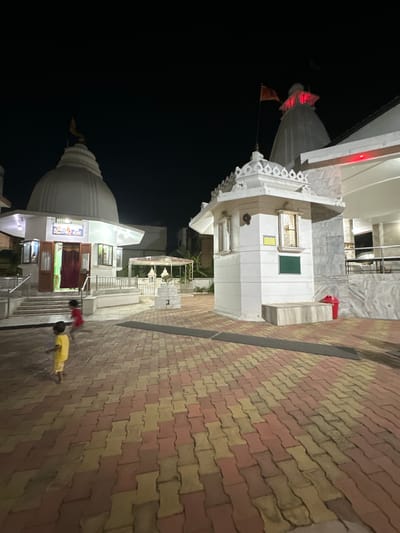 The image captures a nighttime scene at what appears to be a Hindu temple in Vadodara, India. The temple complex features white buildings with a central dome. A pathway, paved with reddish-yellow bricks, leads towards the temple. Two individuals are visible in the image. A child, dressed in yellow, appears to be prostrating on the brick pathway. Another person is seen entering the temple premises. The overall environment is dimly lit, suggesting it is evening or late night.