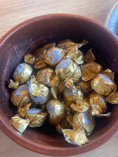 The image displays an overhead shot of a rustic, reddish-brown terracotta bowl filled with numerous individually wrapped chocolates. Each chocolate is encased in shiny golden foil. Visible on several wrappers are white labels bearing the text "GOLDEN LILY" in English and "ЗОЛОТА ЛІЛІЯ" in Cyrillic, indicating the brand "Golden Lily." The chocolates are densely packed within the bowl, with some wrappers appearing slightly crumpled. The bowl itself has an unglazed or roughly finished texture with visible imperfections and variations in its reddish-brown hue. The object is situated on a light-colored wooden surface, displaying distinct grain patterns. A portion of a clear, textured glass object, possibly another dish, is visible in the upper right quadrant of the frame. This scene is documented in Petrivka, Ukraine.