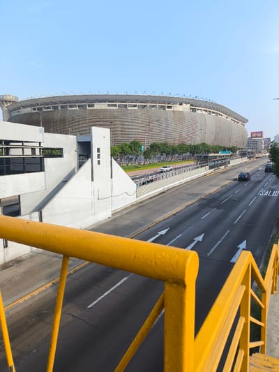 The image shows the Estadio Nacional in La Victoria, Peru, as seen from an elevated pedestrian bridge. The stadium is a large, oval structure with a textured gray exterior and a visible framework on top. In the foreground, the yellow railing of the bridge is prominent. A multi-lane road runs between the bridge and the stadium, with white directional arrows painted on the asphalt and vehicles moving along the road. A bus stop is visible along the side of the road. A building with gray rectangular windows stands on the left side of the road, with a staircase and white concrete features next to it. The sky is blue and clear.