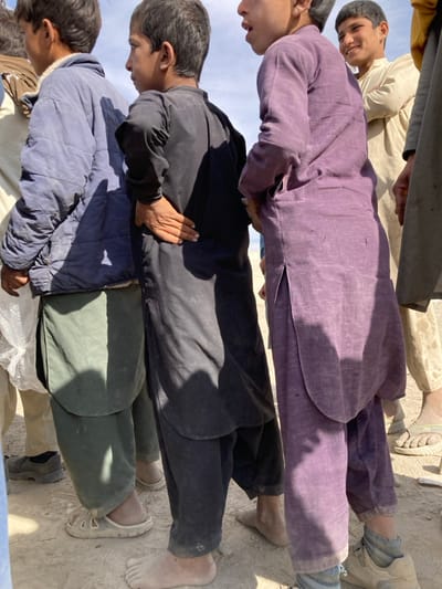 The image depicts a group of at least five young males standing outdoors in Chaman City Tehsil, Pakistan, under bright sunlight. In the foreground, a male on the far left wears a light blue padded jacket over a dark green shalwar kameez, with light-colored sandals visible on his right foot. Adjacent, a young boy in a black shalwar kameez stands barefoot, his right hand resting on his lower back, facing towards the right with his mouth slightly open. To his right, another young boy in a purple shalwar kameez stands, his left hand on his lower back, wearing light-colored athletic shoes with grey socks, also facing right with an open mouth. In the background, to the right of the central figures, a young boy in a light-colored shalwar kameez smiles directly at the viewer, wearing light-colored sandals. Additional partial figures are visible on the extreme left and right. The ground is composed of dry, light-colored earth or sand. Strong sunlight casts distinct shadows, indicating an open, undefined outdoor environment under a clear sky.