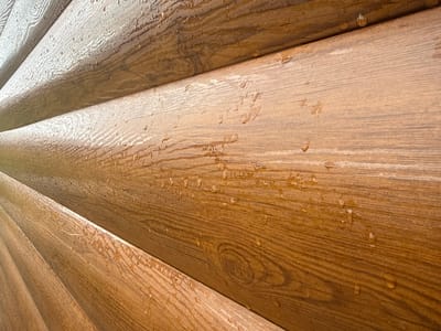 The image displays a close-up, angled view of weathered, brown, wood-like siding, likely on a building. Several rows of siding are visible, with each row appearing to be a single, long plank. The surface of the siding is textured to mimic wood grain, and numerous small water droplets are scattered across it, suggesting recent rain or moisture exposure. The lighting is bright, creating highlights and shadows that accentuate the texture and the water droplets. There are no people or any other discernible objects or actions in the frame. The location is Tuishevo, Russia, and the siding appears to be exterior cladding.