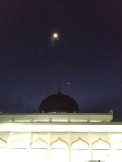 The image captures a serene night scene, possibly in Meuse, Indonesia. The foreground is dominated by the illuminated upper section of a building with arched decorative windows. Above this structure, a large, dark dome, characteristic of a mosque or similar religious building, rises into the deep blue night sky. A slender spire tops the dome.
The sky is dark, indicating nighttime, and features two celestial bodies. The moon is visible as a bright, somewhat diffused orb, partially obscured by wispy clouds, casting a soft glow. A smaller, distinct point of light, likely a planet or a bright star, is also visible to the right of the dome. The lighting on the building's facade suggests artificial illumination, likely for nighttime viewing or security.
There is no discernible text in the image. The overall mood is tranquil and contemplative, emphasizing the quiet beauty of the night sky above the architectural elements. The scene suggests a moment of peace and stillness.