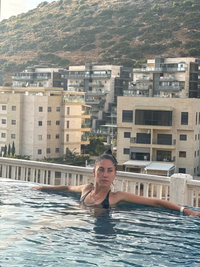 A woman with dark hair pulled back in a bun, wearing a black bikini and a gold necklace, is partially submerged in a swimming pool. She is resting her arms on the edge of the pool. In the background, there are modern apartment buildings with balconies and windows, and a hillside covered in vegetation behind them. The image appears to be taken in Tirat Karmel, Israel, given the architectural style of the buildings and the hilly landscape. The lighting suggests it is daytime, possibly late afternoon.