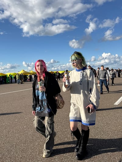In Bolgar, Russia, under a bright blue sky with scattered white clouds, two young individuals walk on an asphalt surface. The person on the left has bright pink hair, wears a black zip-up jacket over a dark graphic t-shirt, and grey cargo pants. They are holding a silver can in their right hand and a patterned bag is slung over their left shoulder. The person on the right has short, lime green hair and is wearing elaborate face makeup. They are dressed in a light-colored, long-sleeved garment with blue trim and dark, lace-up boots. This individual holds a silver can in their left hand and a smartphone in their right. In the background, numerous yellow and green tents are set up, suggesting an outdoor market or festival. Other people are visible walking in the distance, and white lines mark sections of the asphalt ground.