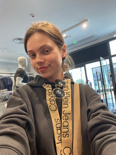 The photograph shows a young woman in what appears to be a clothing store. She is wearing a dark grey sweatshirt with "Calvin Klein Jeans" printed on two tan-colored straps across the chest. Sunglasses are casually placed on the sweatshirt. The background features clothing racks and a mannequin, typical of a retail environment. The lighting suggests an indoor setting, possibly in the city of Marcianise, Italy, given the implied location.