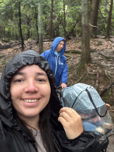 The image depicts two individuals in a dense forest in Nara, Japan, captured from a selfie perspective. In the immediate foreground, a female with dark hair, wearing a black hooded rain jacket with the hood up, smiles directly at the camera. Her face shows freckles, and her clothing appears wet. Behind her, slightly to the right and in the mid-ground, a male wearing a blue hooded rain jacket with the hood up pushes a wheeled transport device. This device, possibly a stroller or cart, is covered by a clear plastic rain cover secured with black straps. Blurred contents, possibly soft items or clothing in blue, pink, and beige, are visible through the transparent cover. The forest ground is covered with damp, fallen leaves, visible tree roots, and earthy debris. Numerous trees with varying bark textures populate the background, indicating a natural, wet, and wooded environment under diffused lighting conditions.