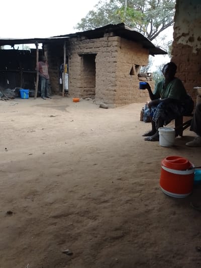 A rural scene features a mud brick house with a makeshift tin roof. A man stands near a blue bucket by the house's open side, while an older woman sits on a wooden stool, holding a blue cup, near the corner of the house. An orange and white water cooler sits on the ground in the foreground. The ground is bare earth. A few trees are visible in the background.