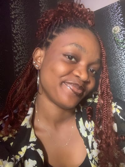 A woman with brown skin smiles, wearing red-toned braids. She is adorned with multiple ear piercings and large silver hoop earrings. A delicate gold necklace graces her neck. She's dressed in a black floral shirt featuring yellow and white flowers. The background is a dark patterned surface, possibly a bed or couch.
