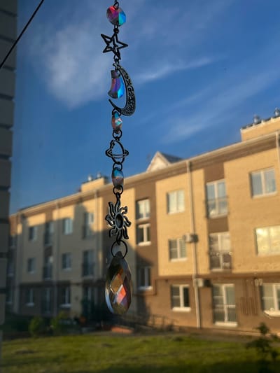 The image shows a suncatcher hanging in front of a window, with a blurry view of a modern apartment building and blue sky with clouds in the background. The suncatcher is made of dark metal chain links, with several iridescent crystal beads and celestial charms. The charms include a star, a crescent moon with intricate filigree, a planet with rings, a sunburst, and a large faceted teardrop crystal at the bottom. The building appears to be made of yellow brick and has multiple windows, some with balconies. The sky is a bright blue with wispy white clouds. The suncatcher and the view through the window suggest a daytime scene. The location of the ЖК «Борисоглебское», Russia is mentioned but not visually depicted in the image itself; it is context provided externally.