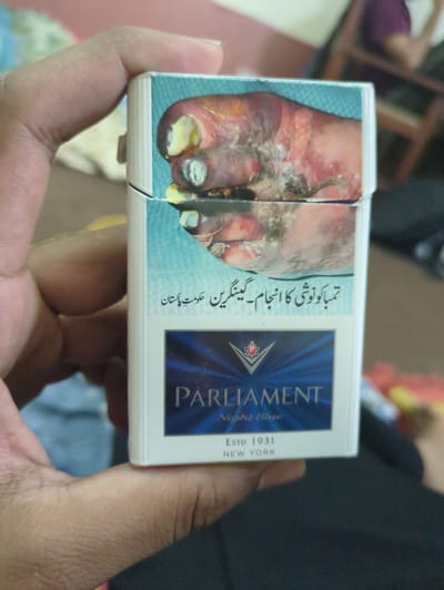 A person's hand is holding a pack of Parliament Night Blue cigarettes. The cigarette pack features a graphic image of gangrenous toes on its front, with text in Urdu below it. The text translates to "The result of tobacco use - gangrene, Government of Pakistan." The background is blurred but appears to be an indoor setting with furniture. The specific location in Abbottabad, Pakistan cannot be determined from the image alone.