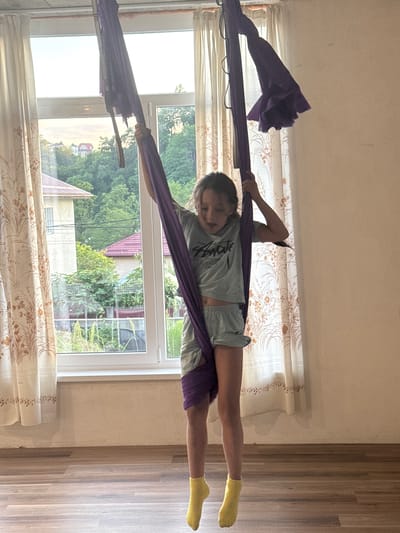 A young person is performing aerial yoga on a purple fabric hammock indoors. The hammock is suspended from above, and the individual is stretched out horizontally, with their legs extended and one hand gripping the fabric for support. They are wearing a light-colored t-shirt, shorts, and yellow socks. The background features a window with a view of trees and houses, suggesting a residential setting, likely in Verkhniy Yurt, Russia. Sunlight is coming through the window, casting shadows on the floor and illuminating the room. The curtains have a floral pattern. The overall mood appears to be one of focus and physical activity. There is visible text on the t-shirt, but it is not clearly legible.