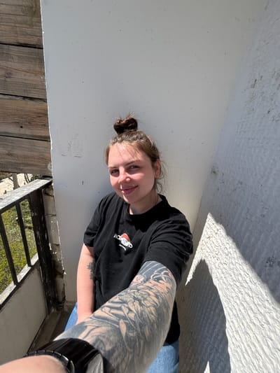 A woman is posing for a photograph, appearing to be outside in what seems to be a partly shaded, outdoor area. The background includes what appears to be a white wall or structure, and below it there's a wooden surface or platform, alongside a metal railing and green vegetation visible through the railing. The woman is wearing a black T-shirt and has a tattoo covering most of her visible arm. There is no explicit visual evidence indicating that this picture was taken in Chornomorskoe, Ukraine.