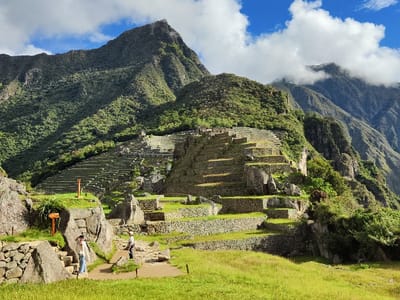Machu Picchu's terraced landscape dominates the scene, set against a backdrop of towering mountains. Lush greenery blankets the slopes, contrasting with the gray stone structures of the ancient city. Two individuals are visible on the lower left, adding a sense of scale. Blue skies and scattered clouds complete the picturesque vista.