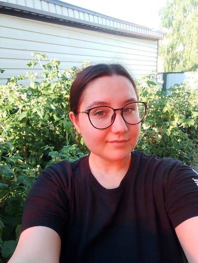 A young woman wearing a black t-shirt and glasses is taking a selfie outdoors. She has dark hair pulled back and is smiling slightly. Behind her, there is a wall of a building and dense green foliage, possibly a garden or bushes. The lighting suggests it is daytime, with sunlight casting some shadows. The scene appears to be a residential area.