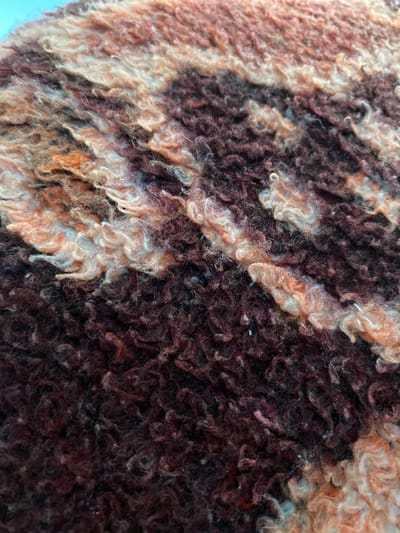 The image displays a close-up, abstract view of a textured material, likely a rug or tapestry, with a pattern of interwoven fibers. The colors are predominantly earthy tones, including deep browns, muted oranges, and creamy whites, creating a striped or banded effect across the surface. The fibers themselves appear thick and curly, giving the material a plush and somewhat shaggy appearance. The lighting suggests a natural light source, highlighting the depth and texture of the fibers. There are no discernible features of the city of Satovcha, Bulgaria, in this image.