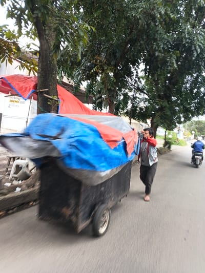 The image displays a daytime street scene in Margahayu, Indonesia, characterized by motion blur suggesting movement. In the foreground, a barefoot male, wearing a grey jacket over a red garment and dark trousers, is actively pushing a large, black, two-wheeled cart. The cart is extensively covered with blue and red tarpaulins. The male holds a red mobile phone to his right ear with his right hand while his left hand is positioned on the cart. On the right side of the paved road, a second individual, dressed in a blue jacket and white helmet, is riding a scooter away from the viewer. The left side of the scene is lined with multiple trees featuring dense green foliage, and makeshift shelters constructed from red tarpaulins. These structures partially obscure a white fence and a visible white sign with black text, including the word "DISEWA" and a phone number. A white plastic stool and some debris are located on the ground near these roadside structures.