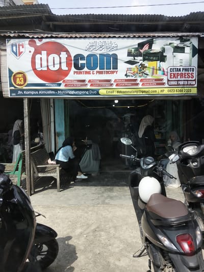 The image shows the exterior of a small shop named "dot com" located at Jln. Kampus Almuslim, Matangglumpangdua, Indonesia. The shop specializes in printing and photocopy services. A banner above the entrance lists services such as Photo Copy, Print A3, Cetak Skripsi, Cetak Undangan, Laminating, and Scan & ATK. It also advertises "CETAK PHOTO EXPRESS" and "SERVICE PRINTER EPSON." Contact information, including an email address (dotcommatang@gmail.com) and phone number (0823 6364 2323), is displayed on the banner.
Inside the shop, several individuals, predominantly women wearing hijabs, are visible. Some are interacting, while others appear to be customers or staff. In the foreground, two motorcycles are parked, suggesting this is a busy area. The building itself appears to be a simple structure with a corrugated metal roof. The overall environment suggests a local business catering to the community's printing and copying needs.