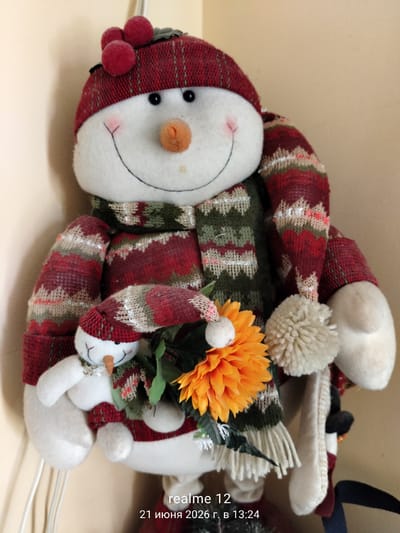 The image is a medium shot of a large, plush snowman decoration, adorned with a red and green knitted hat and scarf. The snowman has black button eyes, a pink blush on its cheeks, and an orange carrot nose. It is holding a smaller, similar-looking snowman toy in its left arm. A bright orange artificial flower is positioned in front of the snowmen, with green foliage. The background is a plain, light-colored wall, likely an indoor setting. In the bottom right corner, there is a watermark indicating "realme 12" and the date "21 июня 2026 г. в 13:24", which translates to "June 21, 2026 at 13:24". The location is identified as Shchyolkovo, Russia.