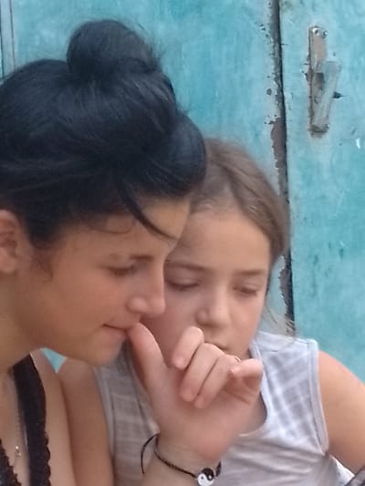 The image shows a close-up of two young girls in Vulcănești, Moldova. The girl on the left, with dark hair pulled back in a bun, is looking down. The girl on the right, with lighter brown hair, is also looking down, with her hand near her mouth, possibly indicating contemplation or shyness. They are positioned in front of a teal-colored, weathered surface that appears to be a wooden door or wall with a metal latch. The lighting suggests it is daytime. The overall scene is intimate and focused on the girls' expressions and proximity to each other.