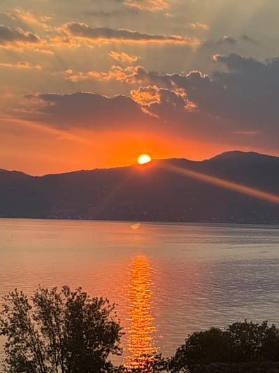 The image depicts a sunset over a body of water, with a silhouetted mountain range in the background. The sun, partially obscured by the mountain crest, casts a warm orange glow across the sky and reflects on the water's surface, creating a shimmering path of light. Wispy clouds are scattered across the sky, catching the sunset colors. In the foreground, dark silhouettes of trees frame the bottom corners of the image. The scene is serene and tranquil, typical of a coastal or lakeside view. This is a picturesque sunset captured from a vantage point overlooking the western shore of Lake Garda, possibly near the town of Garda, Italy, where the mountains meet the lake. There are no people or animals visible in the frame.