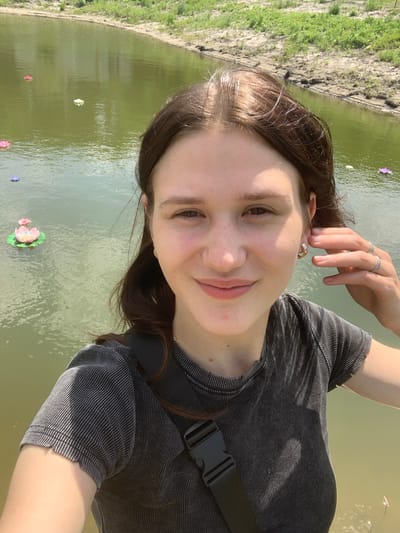 The media file is a selfie of a young woman taken outdoors during the day, likely in the late morning or early afternoon given the bright lighting and shadows. She is smiling and has her hand resting on her head, adorned with two rings. She is wearing a dark, ribbed t-shirt and a black fanny pack across her chest. The background shows a body of calm, green water, possibly a lake or pond, with several decorative objects resembling water lilies floating on the surface. The water is bordered by a muddy bank covered in green grass. The overall mood is cheerful and relaxed, suggesting a leisurely outing. The location is identified as Vasilyevka, Russia. There is no visible text in the image.