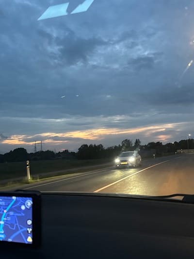 The image captures a forward-facing perspective from inside a vehicle at dusk or early night, on a road identified as being in Saue, Estonia. The sky dominates the upper portion, exhibiting dark blue and grey clouds with a distinct horizontal band of soft orange and pink light along the distant horizon, indicative of a sunset.
In the immediate foreground, the dark-colored dashboard of the recording vehicle is visible. Positioned on the left side of the dashboard is a navigation device, displaying a blue map interface with a bright light-blue route line, various white graphical icons, and a prominent yellow triangular warning symbol. Two light-colored, angular reflections, likely from interior light sources, are visible on the upper part of the windshield.
The two-lane asphalt road extends into the distance, with white dashed lines separating the lanes. A light-colored sedan, possibly white or silver, is approaching in the opposite lane, its headlights fully illuminated and casting a bright glare on the wet road surface. The license plate of this vehicle is present but illegible. The headlights of the recording vehicle illuminate the asphalt directly in front, creating an orange-yellow glow.
To the left of the road, a grassy verge features a white and black reflective marker post. Further in the distance, a dark line of trees and vegetation forms the horizon. Above the treeline, several high-voltage power line towers are clearly visible against the sky. A lone street light is discernible further down the road on the right side. No human figures are explicitly visible, though the presence of the dashboard and navigation device implies a driver within the recording vehicle.