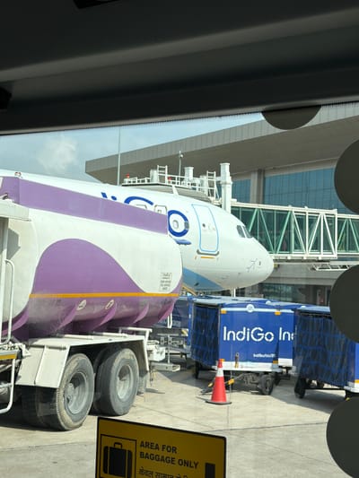 At an airport tarmac in Vikram Nagar, India, a refueling truck with a white and purple tank is parked next to an Indigo Airlines airplane. The truck's wheels are visible, as are several blue cargo containers bearing the Indigo Airlines logo. A jet bridge connects to the airplane, and an airport terminal building is in the background. A yellow sign with a pictogram of a suitcase and text in English and Hindi indicates an area designated for baggage only. The sky is partly cloudy.