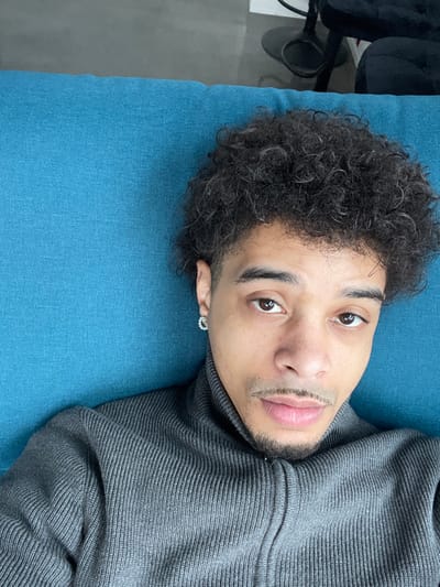 The image, captured in Montigny-le-Bretonneux, France, presents an adult male from an overhead perspective, positioned supine with his head resting on a textured, teal-blue surface. The subject exhibits dark, curly hair with visible gray strands, a neatly trimmed moustache and goatee, and dark brown eyes directed towards the camera. He wears a charcoal gray, ribbed, high-neck sweater featuring a front zipper. A small, circular, possibly jeweled earring is discernible in his left earlobe. The upper right portion of the background, out of focus, reveals components of an indoor environment, including a dark object with a circular base and a section of dark upholstered furniture against a lighter wall.