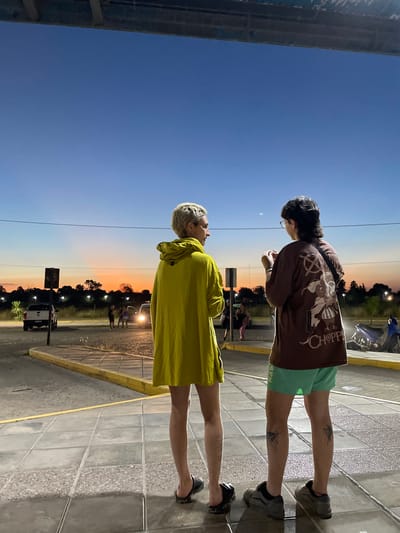 Two people stand on a tiled platform against a backdrop of dusk. The person on the left wears a yellow raincoat and dark sandals. The person on the right wears a brown shirt, green shorts, and gray sneakers. Both figures face away from the camera, looking towards the horizon. The sky fades from blue to orange near the ground. Beyond the platform, a road is lined with trees. A car is visible in the distance.