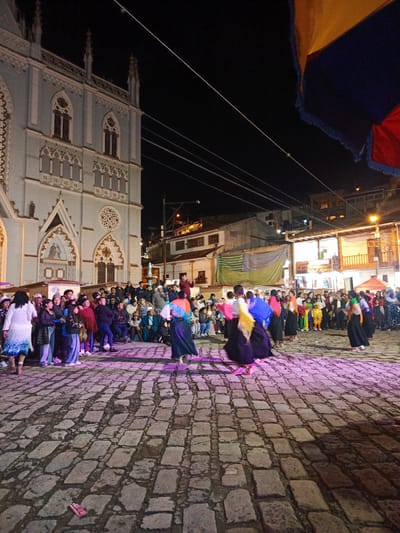 At night in El Cisne, Ecuador, a crowd gathers on a stone-paved plaza in front of a large, ornately decorated church. In the foreground, a group of women in traditional skirts and colorful shawls appear to be dancing. The crowd of onlookers, comprised of people of varying ages, are standing and sitting along the sides of the plaza. Buildings and overhead wires are visible in the background. A portion of a multi-colored flag hangs into the scene from the top right corner.