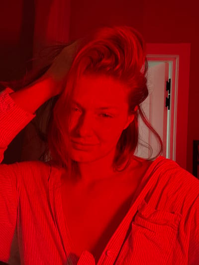 A medium-close-up image depicts a single female individual in an indoor setting, identified by the prompt as the city of Tashevka, Russia. The entire scene is bathed in a strong, monochromatic red artificial light, obscuring the true colors of objects and skin tones.

The subject is positioned centrally, facing directly towards the viewer with a slight, closed-lip smile. Her eyes are open, and she appears to be looking into the camera. Her hair, which appears to be light in color beneath the red light, is styled with volume on top, and her right hand is visible with fingers running through the top-right portion of her hair, slightly lifting it. She is wearing a collared garment, possibly a shirt or blouse, with vertical stripes or a ribbed texture. The garment appears to be partially unbuttoned at the neckline.

In the background, behind and to the right of the subject, a white door with dark hinges is visible, appearing slightly ajar. The wall surrounding the door and the rest of the visible background is uniformly rendered in deep red by the ambient lighting. No other distinct objects or furniture are discernible.