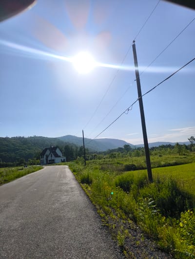 The image depicts a rural landscape on a sunny day, likely in Chelsea, Canada. The foreground is dominated by an asphalt road curving to the left, bordered by lush green grass and wild vegetation. In the midground, a modern white house with a dark roof stands amidst trees and rolling hills. A solitary wooden utility pole with power lines stretches across the frame, adding a touch of infrastructure to the natural setting.

The bright sun is high in the clear blue sky, casting a lens flare that streaks across the image. The sun's intensity suggests it is midday. The surrounding landscape is characterized by gentle, tree-covered hills that recede into the distance, some appearing hazy and layered, indicating a vast expanse. The overall mood is peaceful and serene, typical of a countryside setting bathed in sunlight. No people or animals are visible, and there is no discernible text in the image. The scene evokes a sense of tranquility and natural beauty.