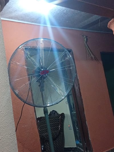 This media file captures an indoor scene in Ukpor, Nigeria, likely during the day given the bright overhead light. The primary subject is a large, metallic oscillating fan, positioned to the left of the frame. Its circular grille, made of thin wires, reflects the strong light. At the center of the fan's grille is a small, red circular emblem. The fan is mounted on a dark green stand, partially visible.
The background consists of an orange-brown textured wall. To the right, a dark wooden doorframe is visible, and within it, a dark interior space is hinted at, possibly another room or closet. Above the wall, a section of the ceiling is visible, appearing textured and possibly stained, with a prominent light source casting intense glare across the top portion of the image.
To the right of the doorframe, a bundle of dry, straw-like material, resembling broom bristles, is hanging from a nail or hook on the wall. Further to the left, a dark, patterned fabric or upholstery is visible, suggesting the presence of furniture, possibly a chair. A black electrical cord for the fan trails down the wall.
A small electrical socket is visible on the wall to the right of the fan, with a small red light illuminated, indicating power. The overall impression is of a functional, somewhat utilitarian indoor space. There is no visible text in the image.