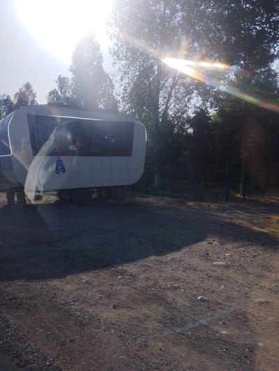 In Balta, Uzbekistan, a white, oblong, trailer-like structure sits on a dirt ground, elevated on four concrete blocks. The structure has large, reflective windows that obscure the interior. A faint reflection of a person's head and shoulders is visible in the windows on the left side. A blue and white sticker, possibly a logo or emblem, is affixed to the side of the trailer. Behind the trailer, a dark fence runs horizontally, separating the area from a wooded background. The sun is bright and positioned high in the sky, casting lens flares of yellow, orange, and pink across the image, particularly over the trees and the trailer. The ground is a mix of dirt and small pebbles.