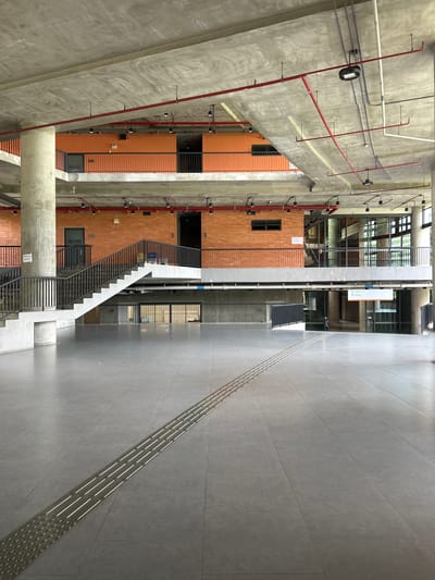The image shows the interior of a modern building with an unfinished, industrial aesthetic. The structure features exposed concrete ceilings, beams, and columns, along with exposed red brick walls. Orange-painted walls are visible on an upper level. Red pipes, likely part of a sprinkler system, run across the ceiling.
The foreground is a large, open, grey tiled floor with tactile paving strips for the visually impaired. A substantial concrete staircase with black metal railings is prominent on the left side of the image, leading up to an upper level. Several doorways and windows are visible on the different levels. On the far right, there's a view through large windows to the outdoors, suggesting the building might be a public space like a school, community center, or transit hub.
The building's architecture suggests a public or institutional use. The presence of tactile paving indicates accessibility for visually impaired individuals. The overall impression is one of a functional and contemporary space, possibly in a public area or a building under construction or renovation. There are no people visible in the image.
This scene does not provide any specific visual cues to confirm its location within Tha Khlong Town Municipality, Thailand.