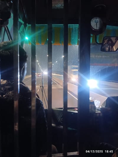 The image presents an interior view from a vehicle at night, looking forward through the windshield, partially obscured by vertical metal bars. The scene is located on a multi-lane road or bridge in Naodoba, Bangladesh. An individual, visible from behind, occupies the front left seat, wearing a patterned shirt. The vehicle's interior includes a dashboard with a round clock and a rearview mirror on the right, reflecting a person. Outside, multiple vehicles are visible on the illuminated road, identified by their bright headlights and red taillights. Streetlights line the thoroughfare, and a distinct green light appears directly ahead. The digital timestamp "04/12/2025 18:45" is present in the bottom right corner.