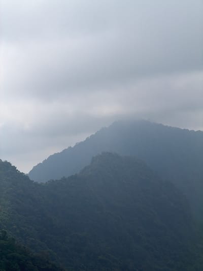 The image shows a panoramic view of lush green mountains under a cloudy sky, likely taken from a high vantage point. The mountains are covered in dense vegetation, with their peaks shrouded in mist. The sky is overcast with thick, gray clouds, suggesting a misty or rainy atmosphere. The overall scene is serene and natural, devoid of any human presence or man-made structures. The location is the mountainous region of Sawan, Indonesia, known for its scenic beauty and natural landscapes.