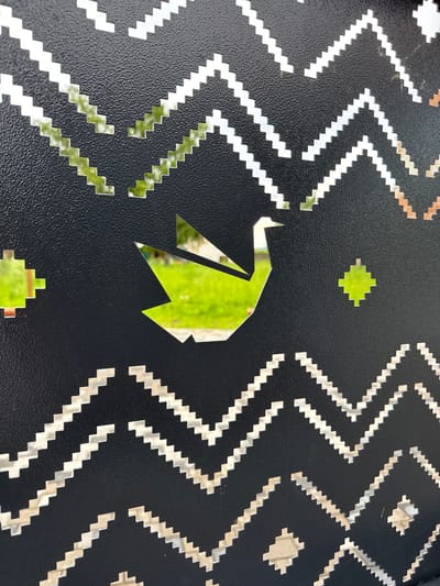 A close-up shot features a black metal panel with a geometric pattern cut out. The pattern consists of jagged, zig-zagging lines and small square shapes. In the center of the panel, a cutout forms the silhouette of an origami bird in flight. Through the cutouts, a brightly lit green lawn and some blurry structures can be seen, suggesting an outdoor environment. The texture of the metal panel is slightly rough. The scene is located in the city of Verkhotor, Russia.