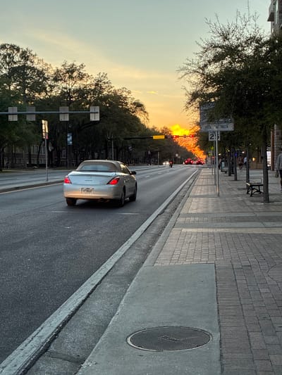 The image captures an urban street scene at sunset in Gainesville, United States of America, viewed from a sidewalk looking east.

In the foreground, a concrete sidewalk section features a circular metal manhole cover with "STORM" etched into its surface. The sidewalk transitions into a patterned brick paver section further down. A concrete curb and gutter separate the sidewalk from the asphalt roadway.

A silver two-door Toyota Solara convertible, with its tan soft top raised, is present in the leftmost lane of the roadway, driving away from the viewer. Its taillights are illuminated.

On the right side of the street, a wide sidewalk is bordered by small, leafy trees. Several indistinct pedestrian figures are visible walking on the sidewalk; some appear to be moving away from the viewer, and one is approaching. Two dark-colored park benches are positioned along the sidewalk. A white sign affixed to a pole reads "BETA PSI THETA" and "SPEED LIMIT 25". Behind the trees and sidewalk, a multi-story building with a modern facade is visible.

On the left side of the street, large, mature trees line the roadway, preceding multi-story buildings. Overhead, a gantry-style traffic light system spans the road, displaying multiple signal heads. A black banner attached to the gantry clearly indicates "GAINESVILLE".

The distant horizon is dominated by the setting sun, casting an intense orange and red glow across the sky and illuminating the far end of the street. Multiple vehicles with illuminated headlights and taillights are visible in the distance, appearing to be stopped at a red traffic light. A motorcycle with a single headlight is also discernible among the distant traffic. The sky transitions from light blue overhead to vivid yellow, orange, and red near the horizon, indicating late afternoon or early evening conditions.