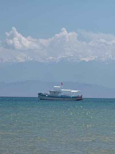 A boat floats on a body of water with snow-capped mountains in the background. The boat is white with a blue stripe along its side and has white canopies. A red flag is visible on the boat. The sky is light blue with fluffy white clouds. The water is a greenish-blue and appears to be choppy. The image appears to be taken from a distance, possibly from the shore or another boat. The scene is set in a natural environment with clear weather. The location is specified as Koshkol', Kyrgyzstan.