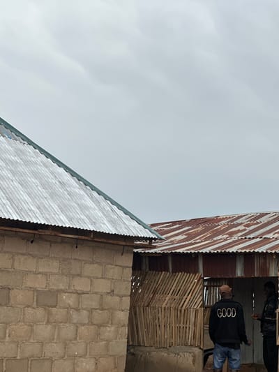In Durumi, Nigeria, two men stand outside a building under a cloudy sky. The man on the left wears a black jacket with the word "GOOD" in gold lettering on the back, a brown baseball cap, and denim shorts. He faces the man on the right, who is wearing a black jacket and dark pants. They appear to be interacting. The building has a brick wall on the left and a corrugated metal roof that is weathered and rusty. To the right of the brick building, there is a section with a wooden slatted wall. The environment suggests a somewhat overcast day, potentially before or after rain.