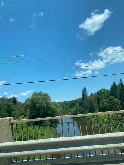 The image shows a view from a bridge over a river, with lush green trees and vegetation lining the banks. The sky is a bright blue with scattered white clouds, and several power lines run horizontally across the top of the frame. The bridge itself has a concrete support on the left and a metal railing. The river appears calm, reflecting the surrounding greenery and sky. The scene suggests a natural, possibly rural or park-like setting within Saint-André-Avellin, Canada.