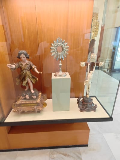In Seville, Spain, within the Museo de Artes y Costumbres Populares, a glass display case showcases religious artifacts. A statue of a child with dark curly hair, wearing a gold-patterned robe, stands on a gilded base adorned with three cherubic heads. To its right, a silver monstrance with a radiating sunburst design is elevated on a rectangular pedestal. Further to the right, a sculpture of Jesus on the cross, carved from ivory, is mounted on a dark, ornate wooden base topped with a skull. The display case is set against a backdrop of polished wooden panels.