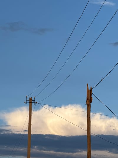 The image captures a view of utility poles and power lines against a twilight sky. On the left, a concrete utility pole stands with insulators and wires. To its right, a wooden utility pole, appearing weathered and partially damaged, is visible. Three black power lines run diagonally across the frame from top right to bottom left. The sky displays layers of clouds; darker, lower clouds are interspersed with lighter, more luminous clouds higher up, suggesting a sunset or sunrise. The scene is set in Baş-Bulak, Kyrgyzstan.