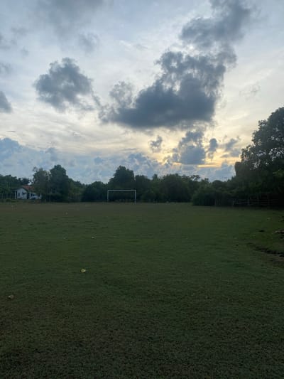 The image depicts an outdoor scene, likely in Tanjongkapai, Indonesia, featuring a large, well-maintained grassy field. In the distance, a white soccer goal is visible, suggesting the field is used for sports. A white house with a red roof is situated to the left of the field, partially obscured by trees. The background is filled with dense green foliage and trees, with some wooden fencing on the right side. The sky above is dramatic, with a mix of dark, heavy clouds and patches of brighter sky, indicating either the early morning or late evening. Scattered on the field are a few small, light-colored objects, potentially debris or fallen leaves. The overall impression is of a rural or semi-rural setting.